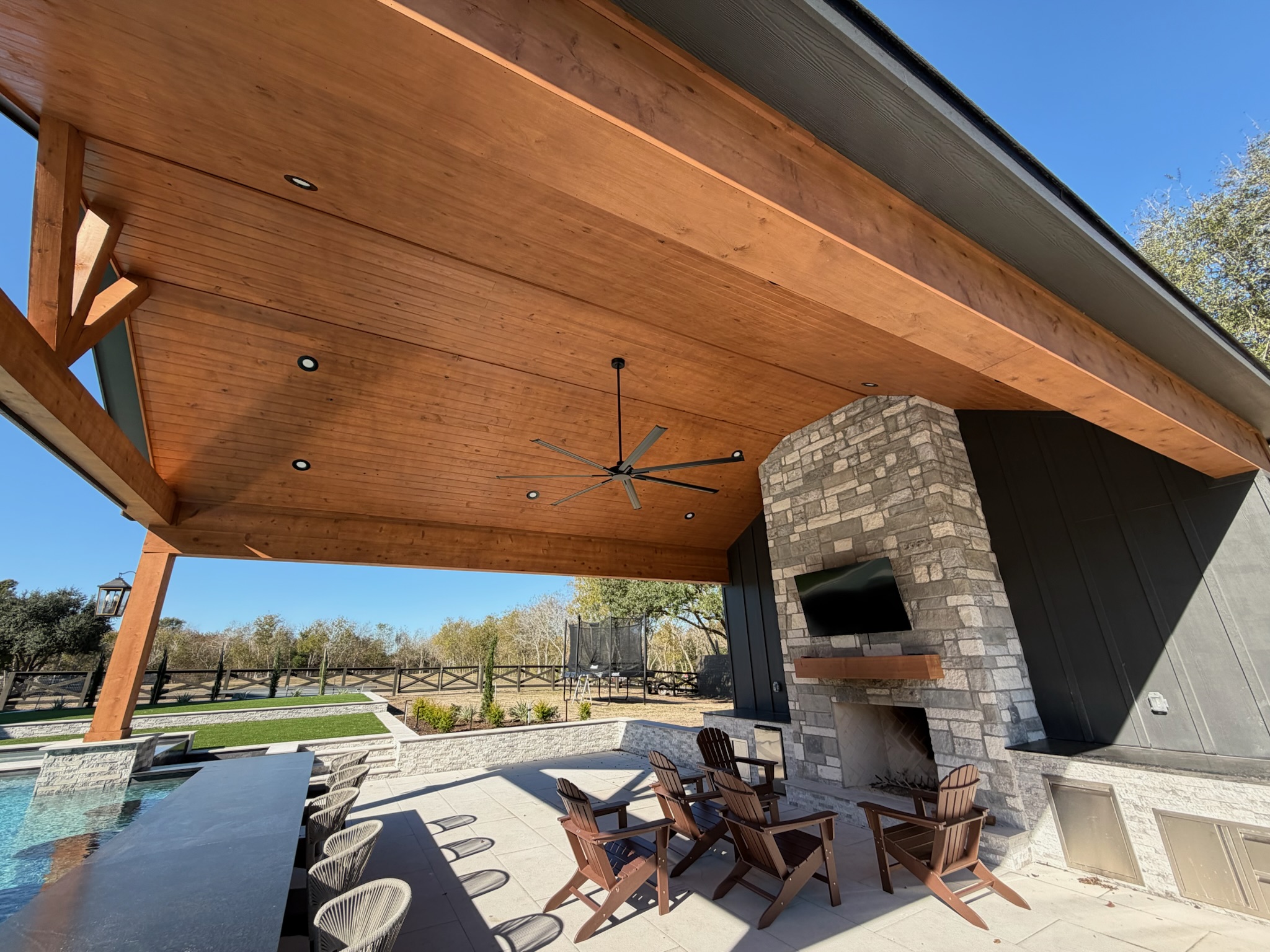 Outdoor patio with wooden ceiling, stone fireplace, pool, and Adirondack chairs